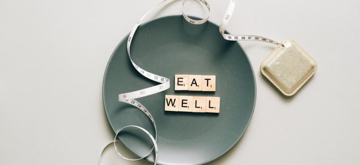 Flat lay of scrabble tiles spelling 'eat well' on a plate with a tape measure, promoting healthy eating.